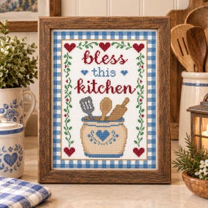 Op de afbeelding: Een ingelijst kruissteek borduurwerk met de tekst "bless this kitchen" in rood en blauw. Het ontwerp toont een beige pot gevuld met een spatel, lepel en deegroller, omringd door rode hartjes en groene rankmotieven.