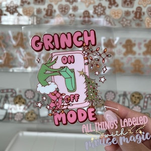 May include: A clear sticker with a pink and green Grinch-themed design. The design features the words "GRINCH ON OFF MODE" with a Grinch hand flipping a light switch. A Christmas tree and string lights are also included.