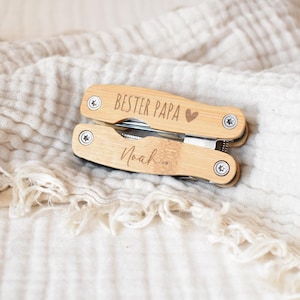 Könnte beinhalten: Ein Holz-Multitool mit den Gravuren "BESTER PAPA" und einem Herz, sowie dem Namen "Noah". Das Werkzeug liegt auf einem weißen, strukturierten Stoff. Ein personalisiertes Geschenk.