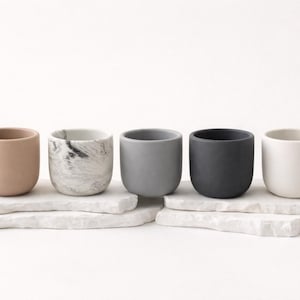 May include: A set of five small, cylindrical cups in varying colours and textures, including a matte tan, a white and black marble pattern, a medium grey, a dark charcoal grey, and a solid white, all arranged in a single row on white stone.