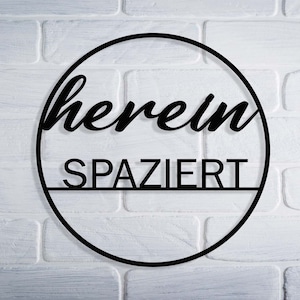 Könnte beinhalten: Schwarzes rundes Schild aus Metall mit dem Text "herein spaziert" an einer weißen Backsteinwand.