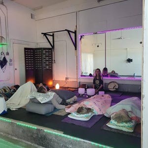 May include: A group of people lying on the floor with blankets and pillows, participating in a sound healing session.  A person is playing crystal singing bowls in the background.