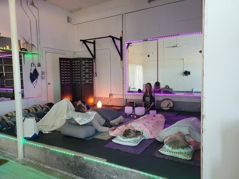 May include: A group of people lying on the floor with blankets and pillows, participating in a sound healing session.  A person is playing crystal singing bowls in the background.