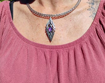 May include: A silver chain necklace with a geometric pendant featuring a purple gemstone. The pendant is shaped like a diamond and has a modern design.