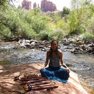 May include: A woman sits cross-legged on a rock by a river, holding a set of wooden flutes. The flutes are decorated with intricate carvings and have a natural wood finish. The woman is wearing a blue skirt and a gray tank top. The background features a rocky landscape and lush greenery.