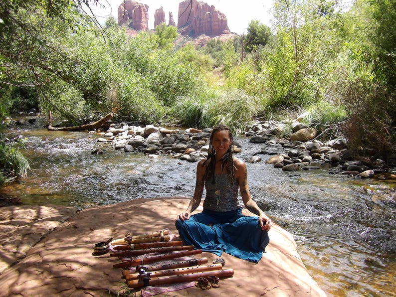 May include: A woman sits cross-legged on a rock by a river, holding a set of wooden flutes. The flutes are decorated with intricate carvings and have a natural wood finish. The woman is wearing a blue skirt and a gray tank top. The background features a rocky landscape and lush greenery.