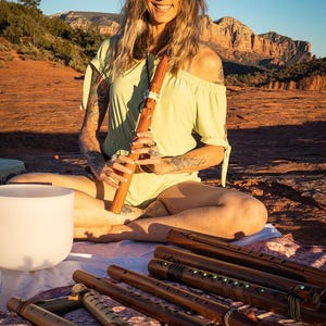 May include: A woman sits outdoors with a wooden flute in her hands. She is wearing a light green off-the-shoulder top and has long brown hair. In front of her are several wooden flutes with intricate designs.