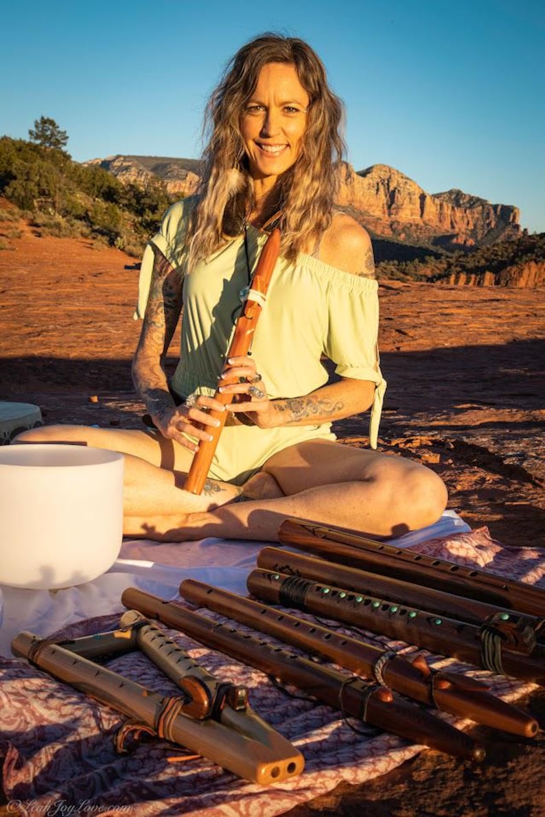 May include: A woman sits outdoors with a wooden flute in her hands. She is wearing a light green off-the-shoulder top and has long brown hair. In front of her are several wooden flutes with intricate designs.