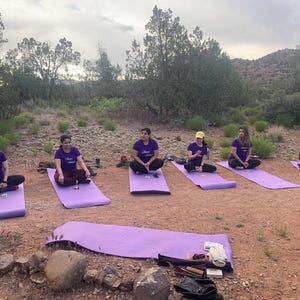 May include: Six people are sitting on purple yoga mats in a circle on a red dirt path. They are all wearing purple shirts with white text. The text on the shirts says "Brave Soul". The people are sitting in a circle in a wooded area with a mountain in the background.