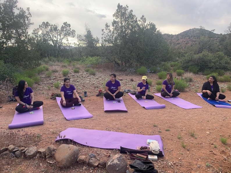 May include: Six people are sitting on purple yoga mats in a circle on a red dirt path. They are all wearing purple shirts with white text. The text on the shirts says "Brave Soul". The people are sitting in a circle in a wooded area with a mountain in the background.