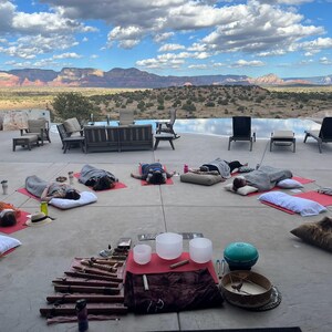 May include: A group of people lying on yoga mats in a relaxed pose on a patio with a view of a mountain range and a swimming pool. There are several singing bowls and other musical instruments in the center of the patio.