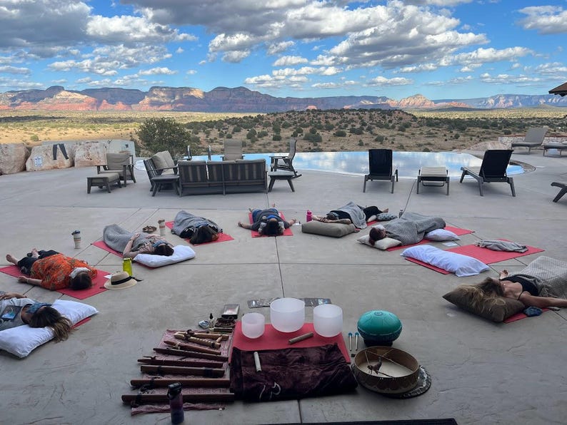 May include: A group of people lying on yoga mats in a relaxed pose on a patio with a view of a mountain range and a swimming pool. There are several singing bowls and other musical instruments in the center of the patio.