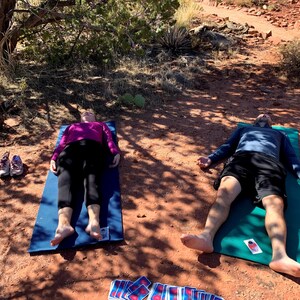 May include: Two people are lying on yoga mats outdoors on a red dirt path. The woman is wearing a purple shirt and black pants. The man is wearing a blue shirt and black shorts. Both have their arms outstretched and eyes closed. There are a few cards scattered on the ground.