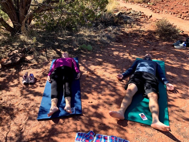 May include: Two people are lying on yoga mats outdoors on a red dirt path. The woman is wearing a purple shirt and black pants. The man is wearing a blue shirt and black shorts. Both have their arms outstretched and eyes closed. There are a few cards scattered on the ground.