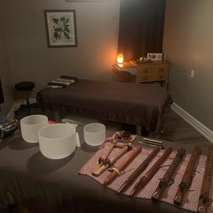 May include: A room with two massage tables, one with a brown blanket and the other with a pink blanket. There are several singing bowls and wooden flutes on the pink blanket. The room is dimly lit with a salt lamp on a wooden table.