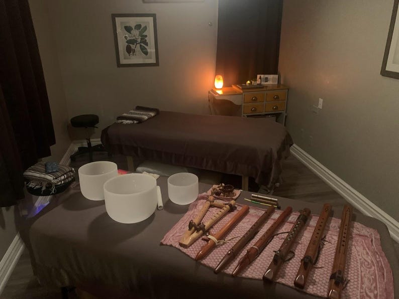 May include: A room with two massage tables, one with a brown blanket and the other with a pink blanket. There are several singing bowls and wooden flutes on the pink blanket. The room is dimly lit with a salt lamp on a wooden table.