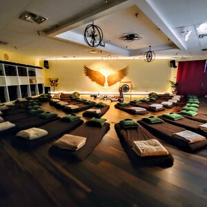 May include: A large room with wooden floors and a circle of brown meditation cushions arranged in a circle. The room has a high ceiling with a large chandelier and a painting of golden wings on the wall.
