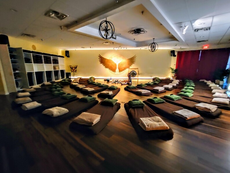 May include: A large room with wooden floors and a circle of brown meditation cushions arranged in a circle. The room has a high ceiling with a large chandelier and a painting of golden wings on the wall.