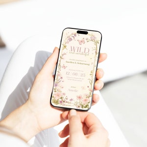 Op de afbeelding: Een smartphonescherm met een digitale verjaardagsuitnodiging met een bloemenrand, vlinders en de tekst WILD and wonderful, Du bist eingeladen zu Amelies 5. Geburtstag, 12 08 25 en deine Amelie in zachte pastelkleuren.