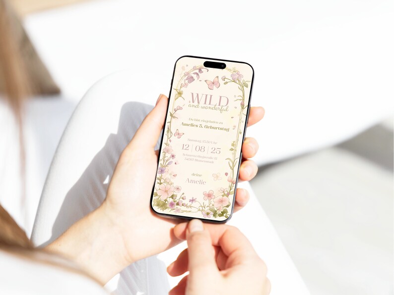 Op de afbeelding: Een smartphonescherm met een digitale verjaardagsuitnodiging met een bloemenrand, vlinders en de tekst WILD and wonderful, Du bist eingeladen zu Amelies 5. Geburtstag, 12 08 25 en deine Amelie in zachte pastelkleuren.
