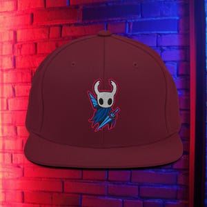 Puede incluir: Gorra de béisbol granate con visera plana. La gorra presenta un diseño bordado de un personaje blanco, azul y negro sosteniendo una espada. El fondo es una pared de ladrillos con iluminación roja y azul.