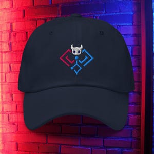Puede incluir: Gorra de béisbol azul marino con un diseño bordado. El diseño presenta una figura blanca con cuernos dentro de una forma geométrica roja y azul. La gorra tiene una visera curva y un cierre de correa de tela.