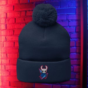 May include: Dark navy beanie hat with a large pom-pom. The hat has an embroidered emblem of a character with a cape and sword in red, blue, and white. The beanie is made of a soft, knitted material.