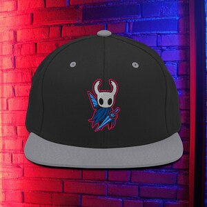 Puede incluir: Gorra de béisbol negra y gris con visera plana y diseño bordado. El diseño es un personaje blanco con una capa azul y una espada azul. La gorra tiene una visera gris y un botón gris en la parte superior.