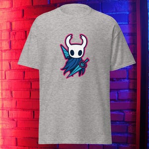May include: A grey t-shirt with a cartoon character design. The character has a white head, blue cape, and a sword, outlined in pink. The shirt is made of a soft material.