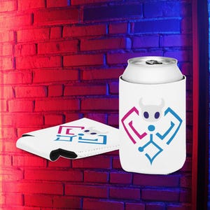 Puede incluir: Enfriador de latas blanco con un diseño de toro geométrico en azul y magenta. Una lata plateada está dentro del enfriador. Otro enfriador plano con el mismo diseño está al lado. El fondo es una pared de ladrillos rojos.
