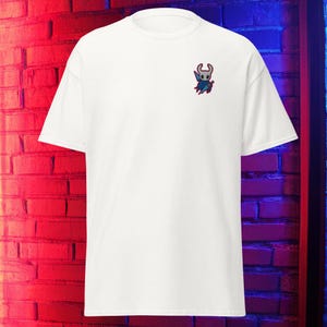 Puede incluir: Una camiseta blanca con un pequeño diseño bordado de un personaje con un casco con cuernos y una capa. El diseño es en azul, rojo y blanco. La camiseta tiene mangas cortas y cuello redondo.