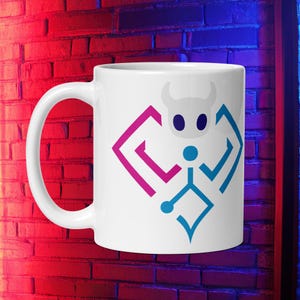 Puede incluir: Taza de cerámica blanca con un diseño geométrico en rosa y azul. El diseño incluye una figura estilizada con cabeza con cuernos, una forma de diamante y un cuerpo similar a un hombre de palo. La taza tiene un asa curva.