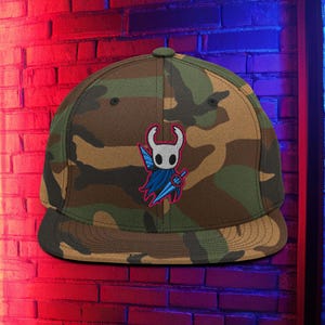 May include: A camouflage-patterned flat-brimmed cap featuring a cartoon character with a white face, blue cape, and a sword. The cap has a brown, green, and tan camouflage design. The character is embroidered on the front.