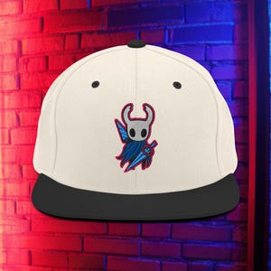 Puede incluir: Gorra blanca y negra de ala plana con un diseño bordado de un personaje con cabeza blanca, capa azul y espada azul y plateada. La gorra tiene una visera negra y un botón negro en la parte superior.
