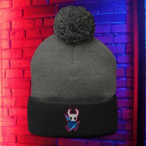 May include: A grey beanie hat with a black brim and a grey pom-pom. The black brim features a colourful embroidered design of a character with a blue cape and a sword. The hat is displayed against a brick wall with red and blue lighting.
