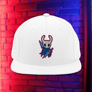 Puede incluir: Gorra de béisbol blanca con visera plana, bordada con un diseño de personaje. El personaje tiene una cabeza blanca, cuernos rojos, capa azul y una espada azul. La gorra tiene una corona estructurada y una visera curva. Fondo de pared de ladrillo con iluminación roja y azul.