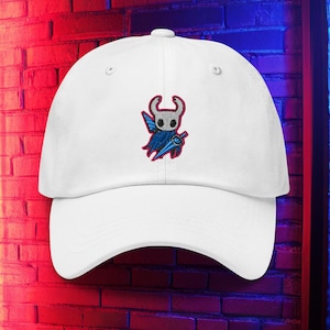 May include: White baseball cap with a curved peak. The cap has an embroidered design of a character with a white head, red horns, blue cape, and a blue sword. The cap has a classic six-panel design with a curved visor.