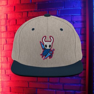 May include: A beige baseball cap with a navy blue peak, featuring an embroidered character design with a white head, blue cape, and a blue sword. The cap has a flat peak and a button on top.
