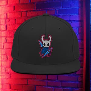 Puede incluir: Gorra negra de ala plana con un diseño bordado de un personaje blanco, azul y rojo sosteniendo una espada. La gorra tiene una corona estructurada y una visera plana. El diseño está centrado en la parte delantera.