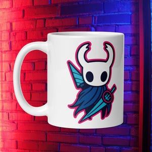 May include: White ceramic mug with a cartoon character design. The character has a white head, blue cape, and a teal sword, outlined in pink. The mug has a large handle. The background is a brick wall.