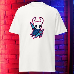 Puede incluir: Camiseta blanca con un diseño de personaje de dibujos animados. El personaje tiene una cabeza blanca, una capa azul y una espada, todo delineado en rosa. La camiseta está hecha de un material suave.