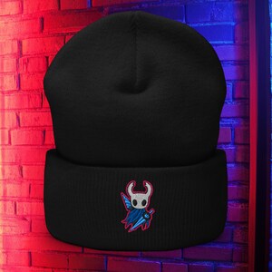 May include: A black beanie hat with a folded brim. The hat features an embroidered design of a character with a white face, blue cape, and a sword. The background is a brick wall with red and blue lighting.