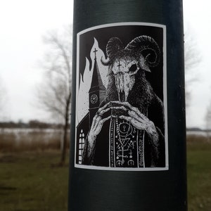 May include: Black and white sticker of a horned demon with a skull-like face, holding a book with the text "IN NOMINE DEI NOSTRI SATANAI" and "Gnosis Occult" in a gothic font. The demon is standing in front of a church with flames behind it.