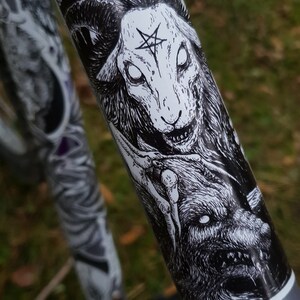 May include: Black and white sticker of a demonic sheep with a pentagram on its forehead. The sheep is snarling and has large, white eyes. The sticker is on a gray bike frame.
