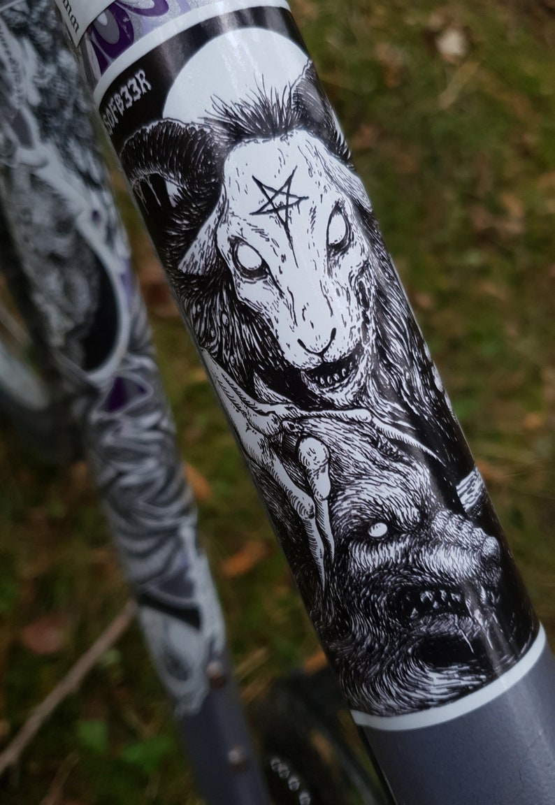 May include: Black and white sticker of a demonic sheep with a pentagram on its forehead. The sheep is snarling and has large, white eyes. The sticker is on a gray bike frame.