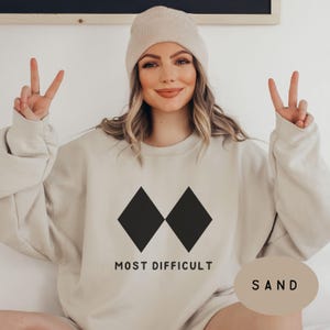 May include: A sand-colored sweatshirt with the words "MOST DIFFICULT" printed below two black diamond shapes. The word "SAND" is printed on a sand-colored oval. A person is wearing a sand-colored beanie.