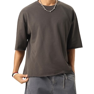 Può includere: Una t-shirt grigio antracite a maniche corte con scollo rotondo e bordi grezzi non rifiniti sulle maniche e sull'orlo inferiore. La maglia presenta una vestibilità ampia e squadrata, abbinata a pantaloni cargo grigio scuro con coulisse.