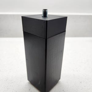 May include: A black wooden furniture leg with a metal screw thread. The leg has a square top and a tapered shape. The black paint has a matte finish. The furniture leg is designed for easy installation, and is suitable for various furniture items.