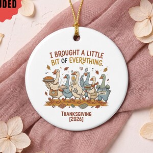 May include: A round white ceramic ornament hanging from a gold string. The center features a colorful illustration of five geese carrying Thanksgiving food with the text I BROUGHT A LITTLE BIT OF EVERYTHING and THANKSGIVING [2026] printed below.