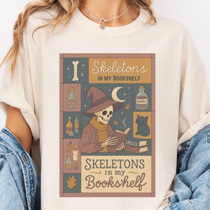 May include: Cream-colored t-shirt featuring a rectangular graphic with the text "Skeletons in my Bookshelf." The graphic depicts a skeleton wearing a witch hat, reading a book, with potion bottles and a black cat. The shirt has short sleeves.